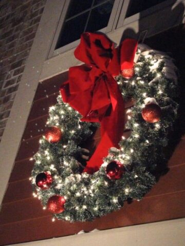 Christmas wreath hung on a house with red ornaments and white lights.