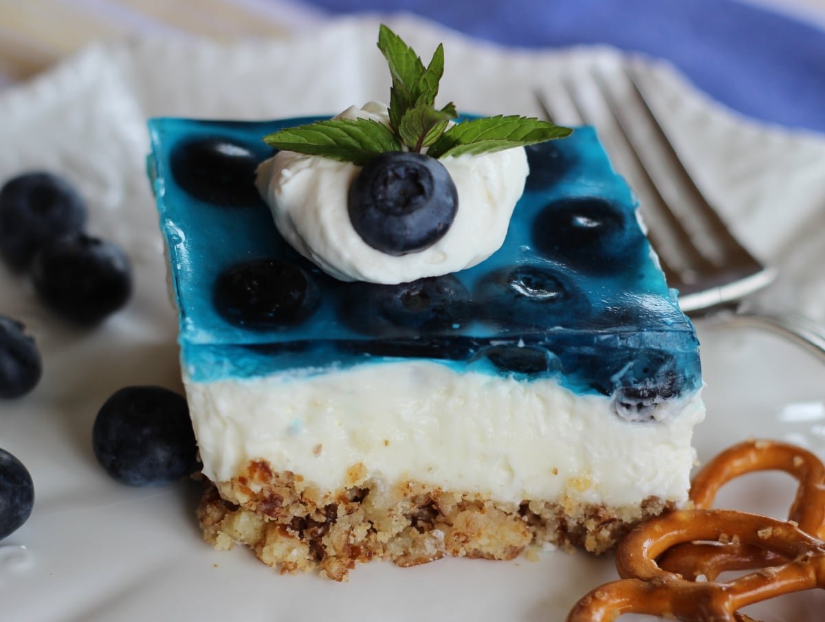 a piece of blueberry pretzel dessert with a base layer of pretzel crumbs, then a light cloud layer of cream cheese and whipped cream and on top blueberries in blueberry jello.