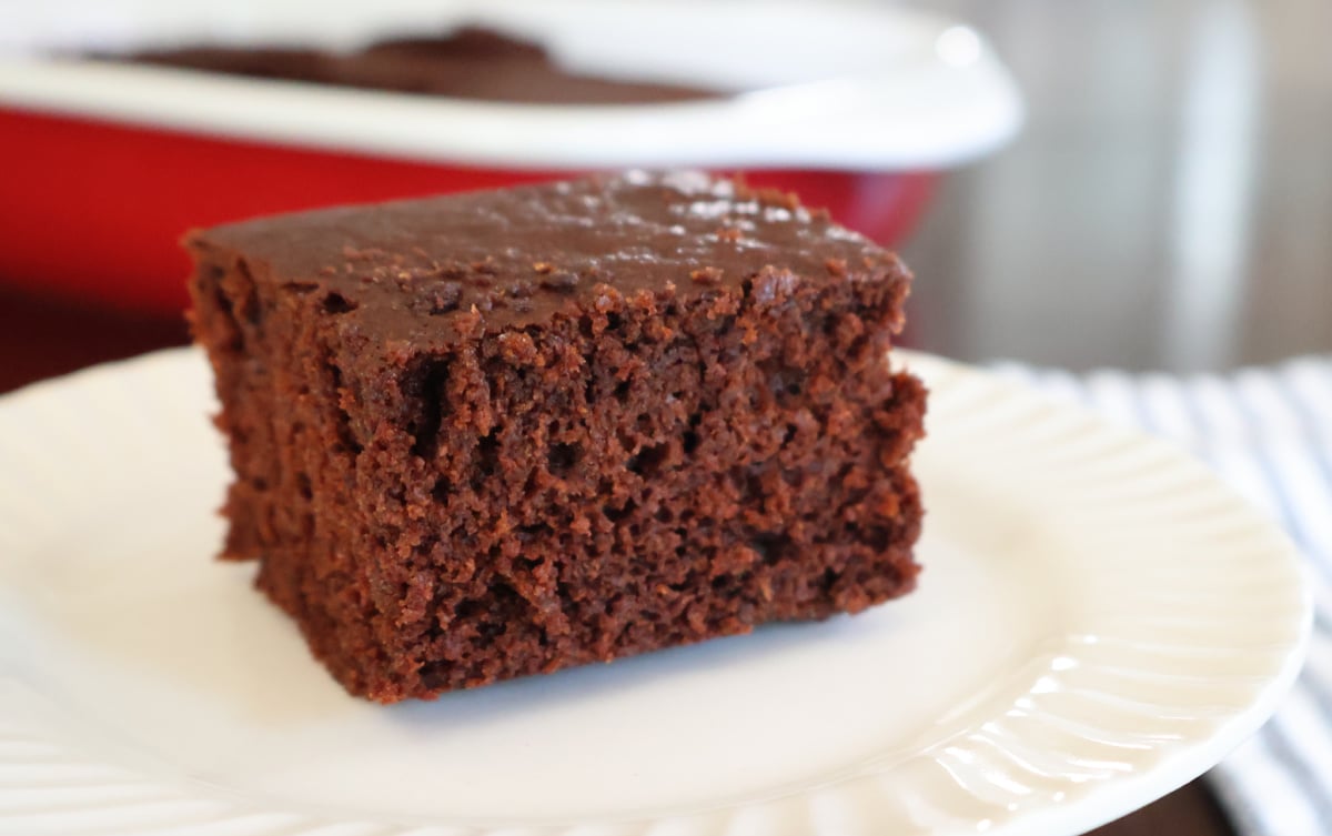 A square piece of brownie cake on a white bowl with the red baking dish in the background.