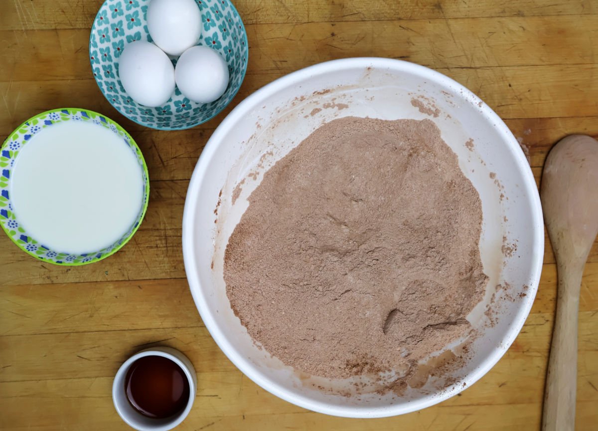 dry ingredients for brownie cake have been stirred together and some of the wet ingredients like eggs, milk and vanilla are ready to be added to the bowl.