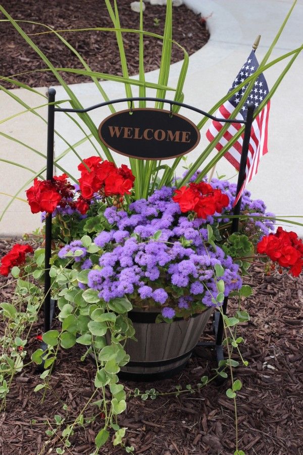 A welcome sign over a half barrel containing red and purple flowers and green leafy plant