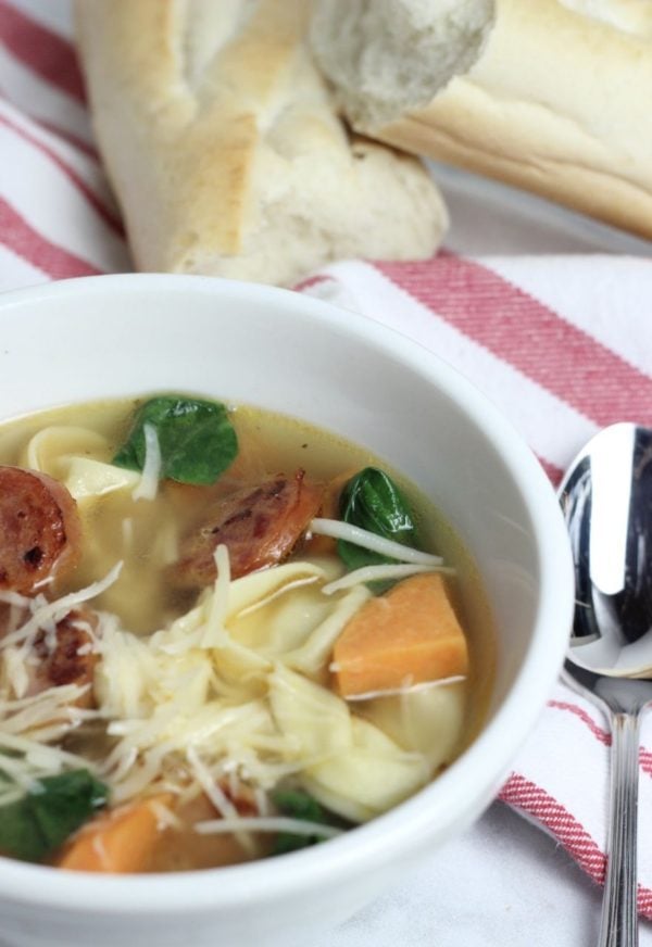 a bowl of soup with sliced brats, ravioli, cut up squash and spinach.