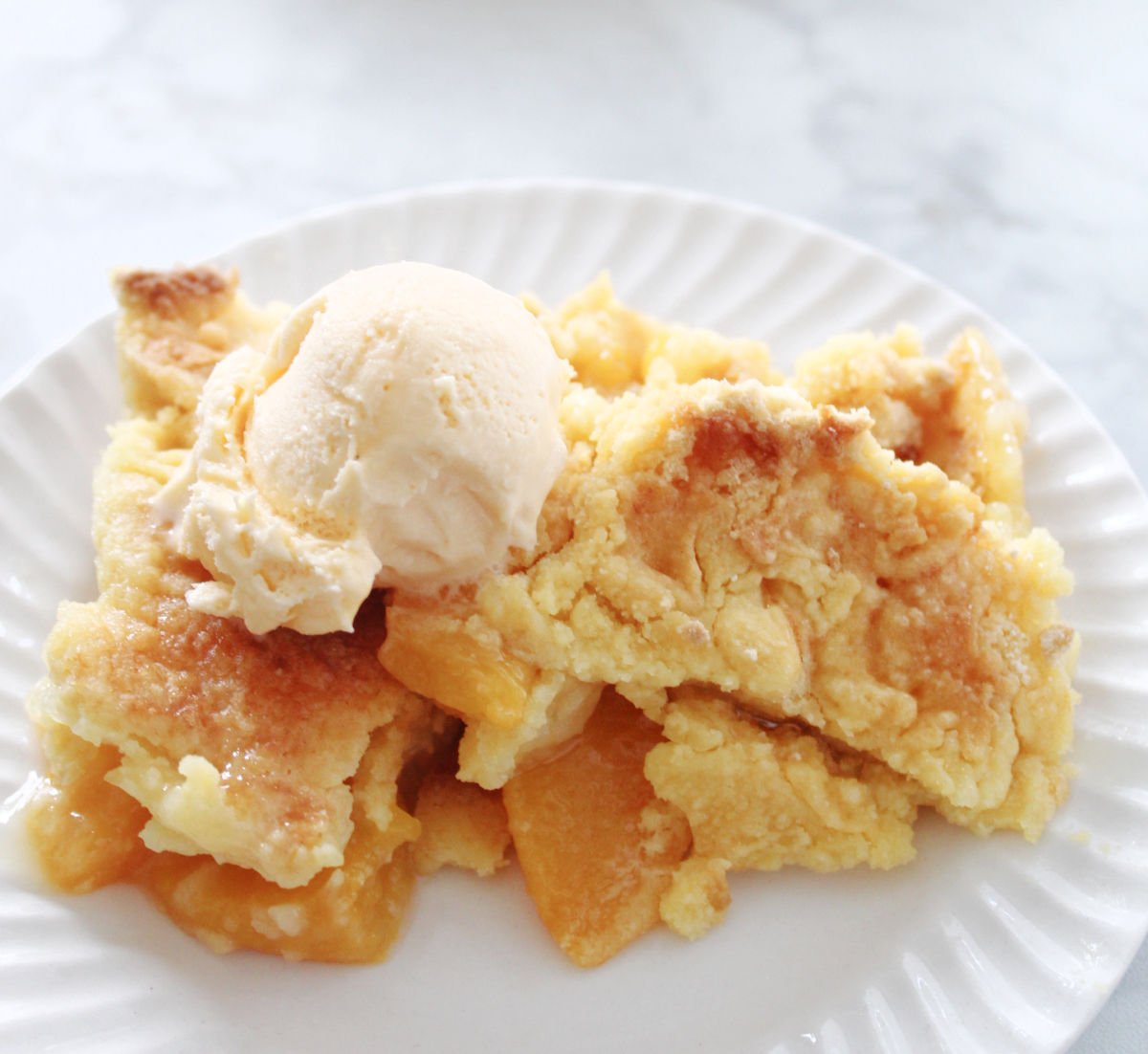 a serving of peach cobbler with a crispy topping and peaches showing. Served on a white plate.