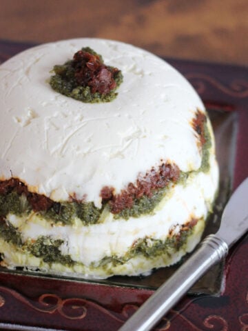 Flavorful and colorful layers of green pesto with red sundried tomatos between a cream cheese layer base. Layers are added to a bowl to form a rounded shape.
