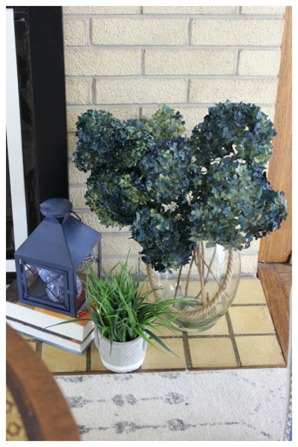 A display beside the fireplace containing blue annabelle hydrangeas, a blue lamp and a small green leafy plant.