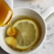 lemon ginger tea with a slice of lemon and slices of ginger in a white cup. Honey is drizzling into the cup.