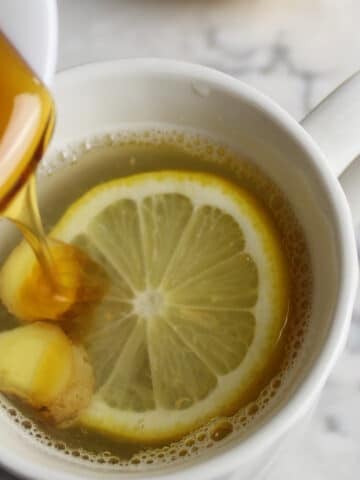 lemon ginger tea with a slice of lemon and slices of ginger in a white cup. Honey is drizzling into the cup.