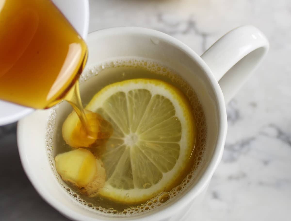lemon ginger tea with a slice of lemon and slices of ginger in a white cup.  Honey is drizzling into the cup.