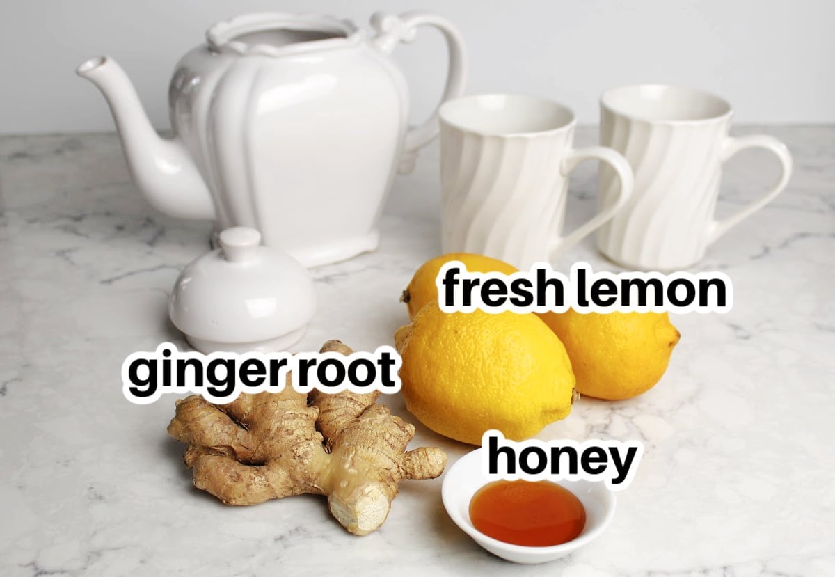 ingredients for lemon ginger tea including fresh lemon, ginger root and honey.  White teapot and two teacups in the background. 