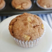 An apple spice muffin with pieces of apple showing. On a white plate with three muffins visible in a muffin tin behind the main muffin.