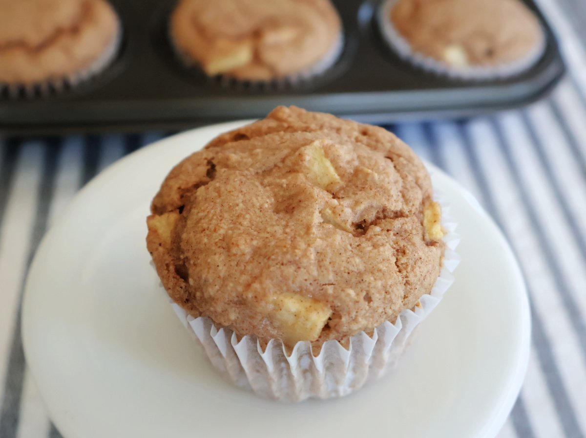 An apple spice muffin with pieces of apple showing. On a white plate with three muffins visible in a muffin tin behind the main muffin.