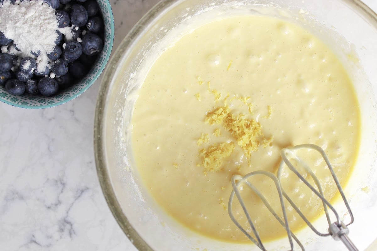 batter for blueberry muffins that has freshly grated lemon on top of batter.  Mixer beaters are ready to go into batter.  Blueberries are in a small bowl to the left of large batter bowl (with cake mix on the blueberries).