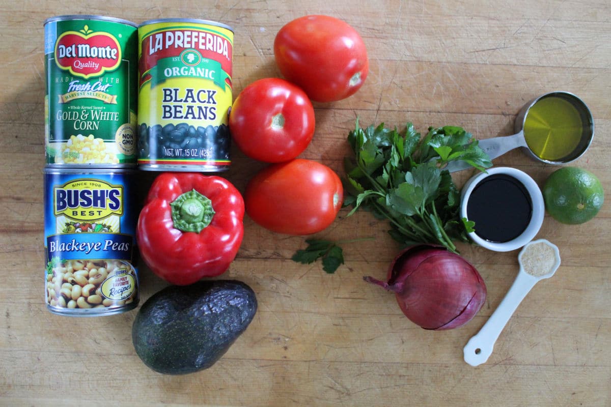 Ingredients for cowboy caviar including black beans, buckeye peas, corn, bell pepper, tomato, avocado, parsley, avacodo oil, salsalmic vinegar, lime, onion and garlic salt.