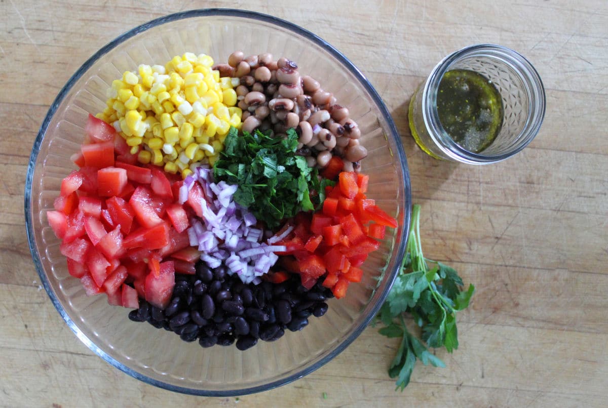 Ingredients for cowboy caviar including corn, black beans buckeye peas, chopped tomato, bell pepper, red onion, parsley and avacado.  Dressing in a mason jar is ready to be added.