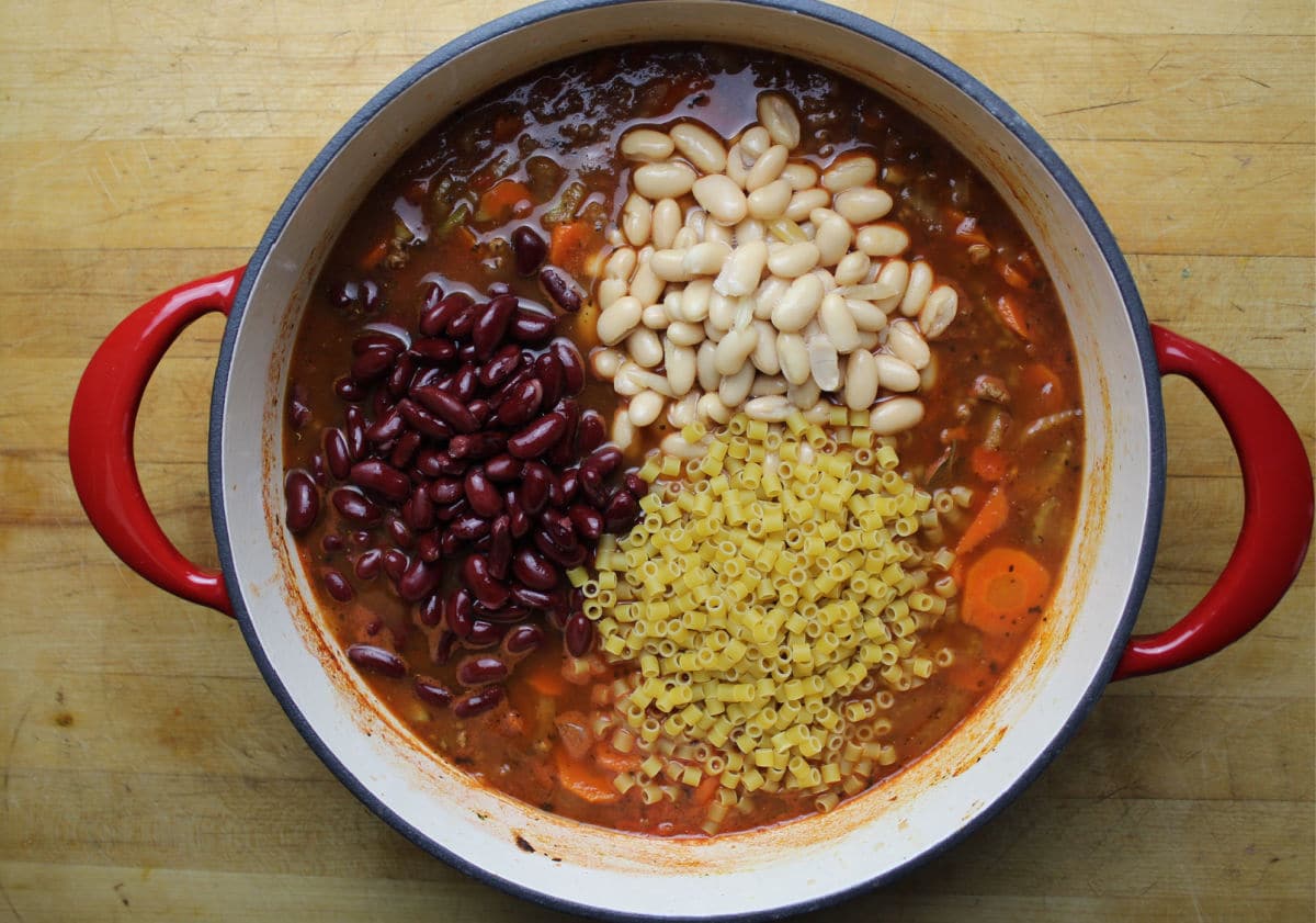 Beans and pasta added to soup in a red pot.