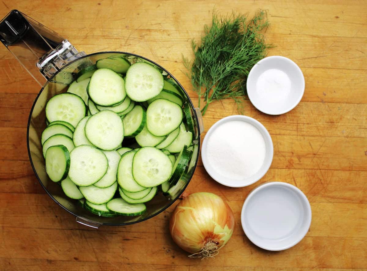 sliced cucumbers with an onion, fresh dill, sugar and vinegar beside the container of cucumber slices.