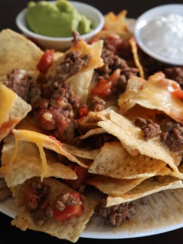 plate with ground beef nachos with melted cheese. guacamole and sour cream served in small containers to dip.