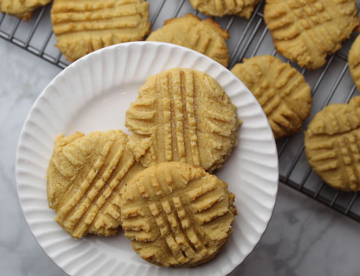 Three peanut butter cookies on a white plate with other cookies on a cooling rack in the background.