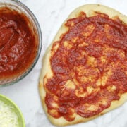 pizza sauce in a small bowl and beside that a piece of oval flatbread covered in tomato sauce.