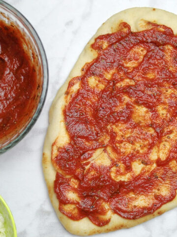 pizza sauce in a small bowl and beside that a piece of oval flatbread covered in tomato sauce.