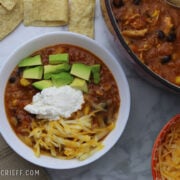 A white bowl full of chicken tortilla soup garnished with cut up avocado, Monterey Jack cheese and sour cream in the middle. The pot of soup and a bowl of shredded cheese is to the right of the soup bowl.