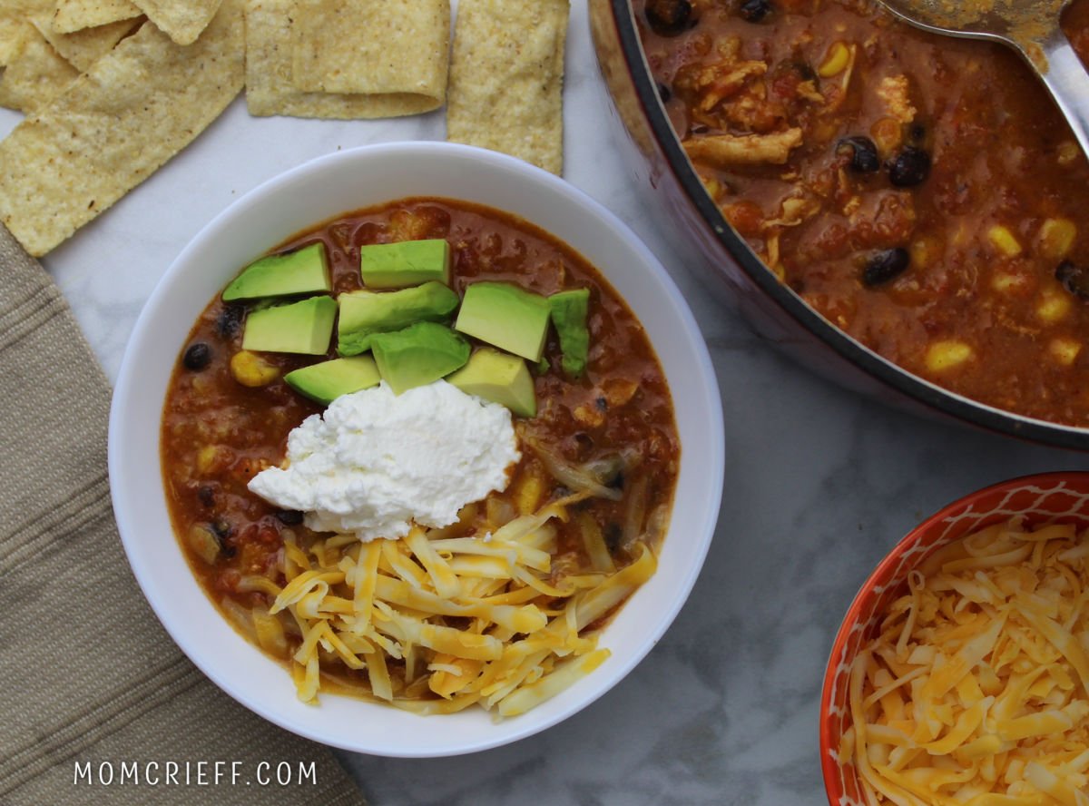 A white bowl full of chicken tortilla soup garnished with cut up avocado, Monterey Jack cheese and sour cream in the middle. The pot of soup and a bowl of shredded cheese is to the right of the soup bowl.