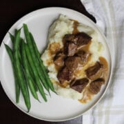 Beef piecen on mashed potatos with gravy on a white plate. Green beens are to the left of the beef and potatos.