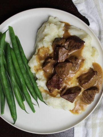Beef piecen on mashed potatos with gravy on a white plate. Green beens are to the left of the beef and potatos.