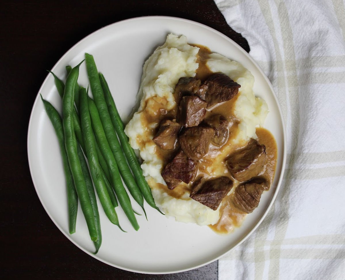 Beef piecen on mashed potatos with gravy on a white plate. Green beens are to the left of the beef and potatos.