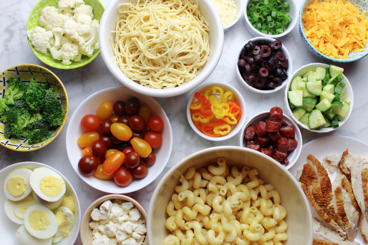 pasta bar ingredients including cooked spaghetti, spiral pasta, tomatos, cucumbers, steamed broccoli, hard boiled eggs, feta cheese, cut up rotisserie chicken, peppers.