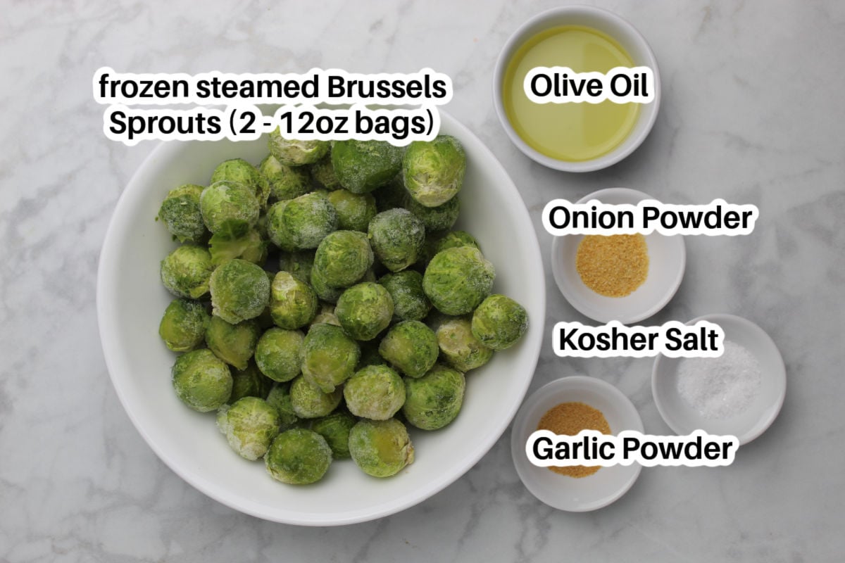 A medium bowl with frozen Brussels sprouts, small white dishes containing olive oil, onion powder, kosher salt and garlic powder.