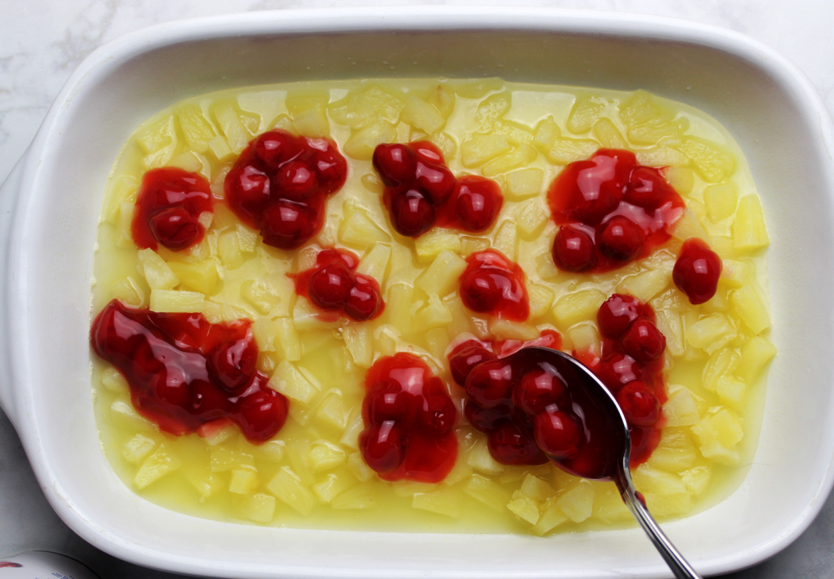 spooning cherry pie layers over pineapple for the cherry pineapple dump cake recipe