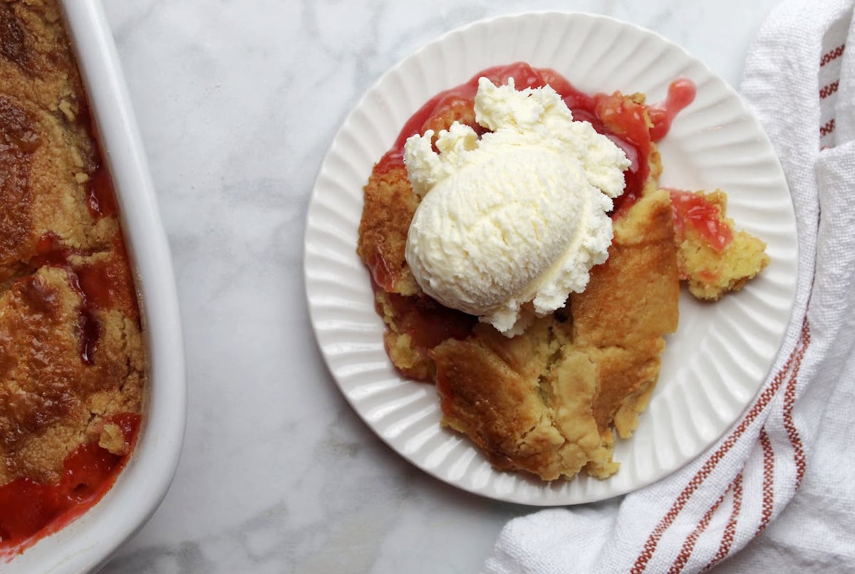 Cherry pineapple dump cake served on a white plate with a big scoop of vanilla ice cream. A part of the casserole can be seen with the baked dump cake on the left side of the image.