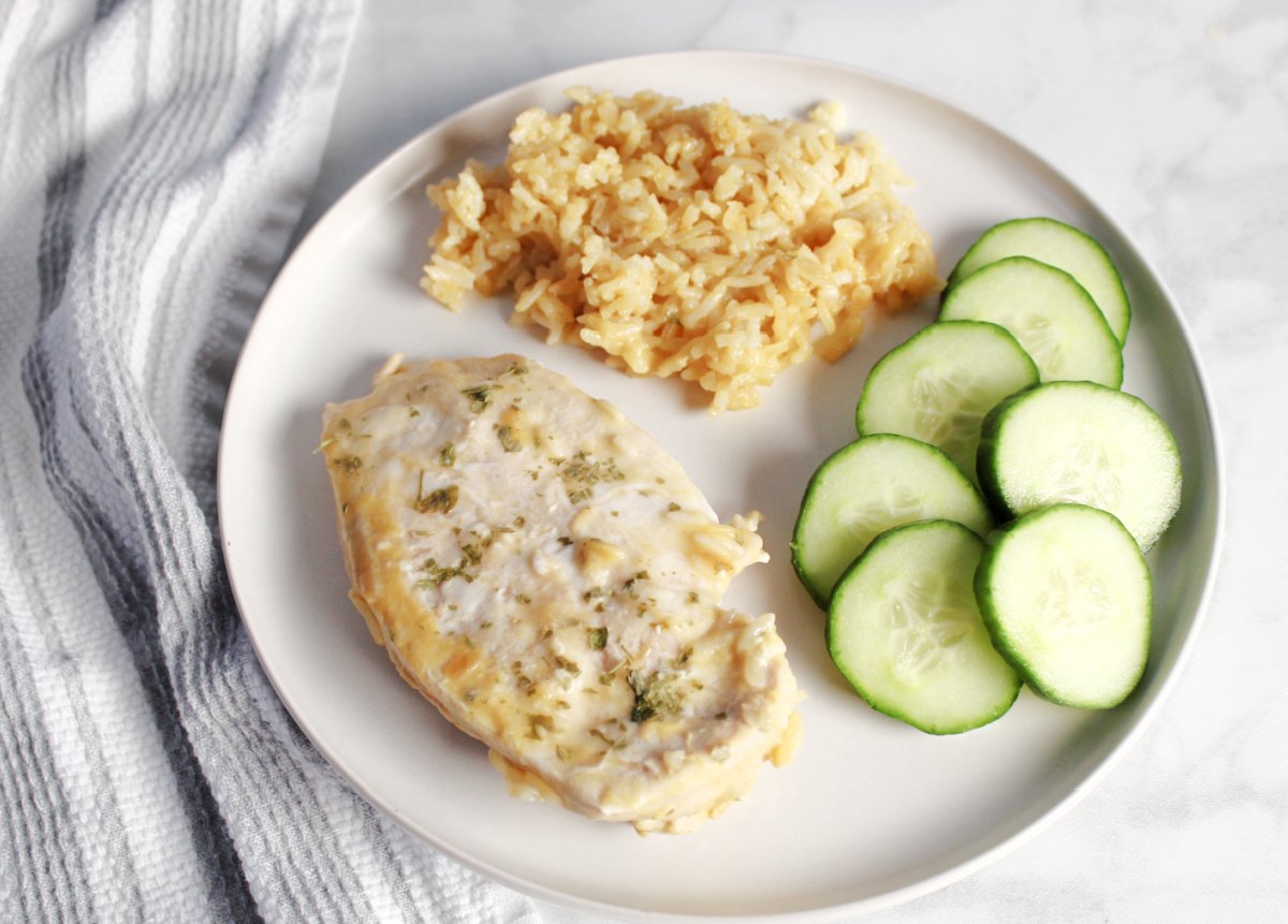 A piece of no peek chicken with seasoning, a serving of flavored rice and some fresh cucumbers as a side on a white plate.