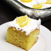 A piece of yellow lemon poke cake with whipped topping and a piece of lemon as a garnish on a white plate. The pan with the rest of the lemon poke cake in behind and all the cut pieces are garnished with ¼ of a lemon slice.