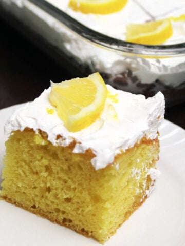A piece of yellow lemon poke cake with whipped topping and a piece of lemon as a garnish on a white plate. The pan with the rest of the lemon poke cake in behind and all the cut pieces are garnished with 1/4 of a lemon slice.
