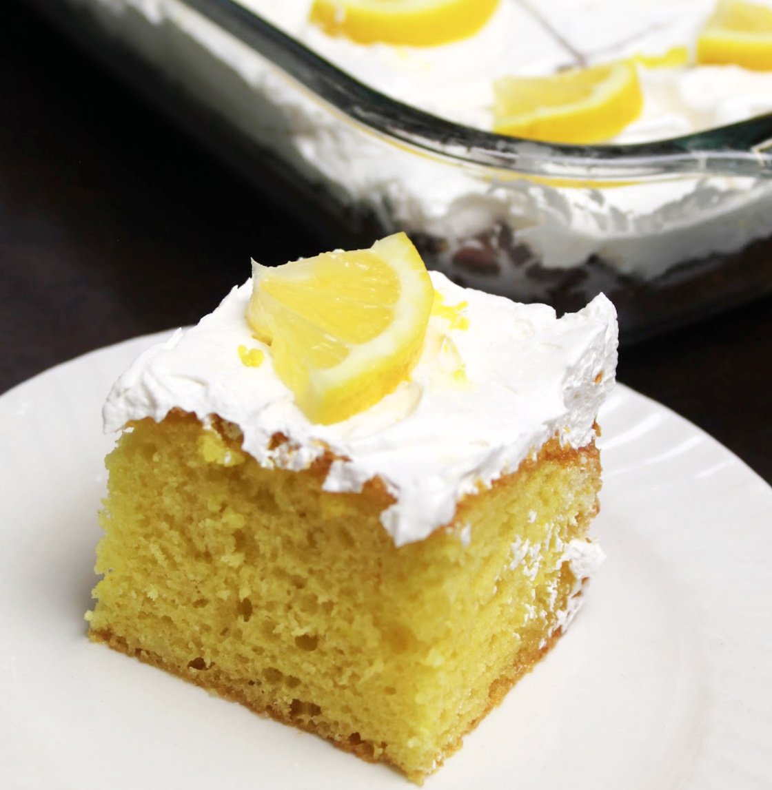 A piece of yellow lemon poke cake with whipped topping and a piece of lemon as a garnish on a white plate.  The pan with the rest of the lemon poke cake in behind and all the cut pieces are garnished with ¼ of a lemon slice.