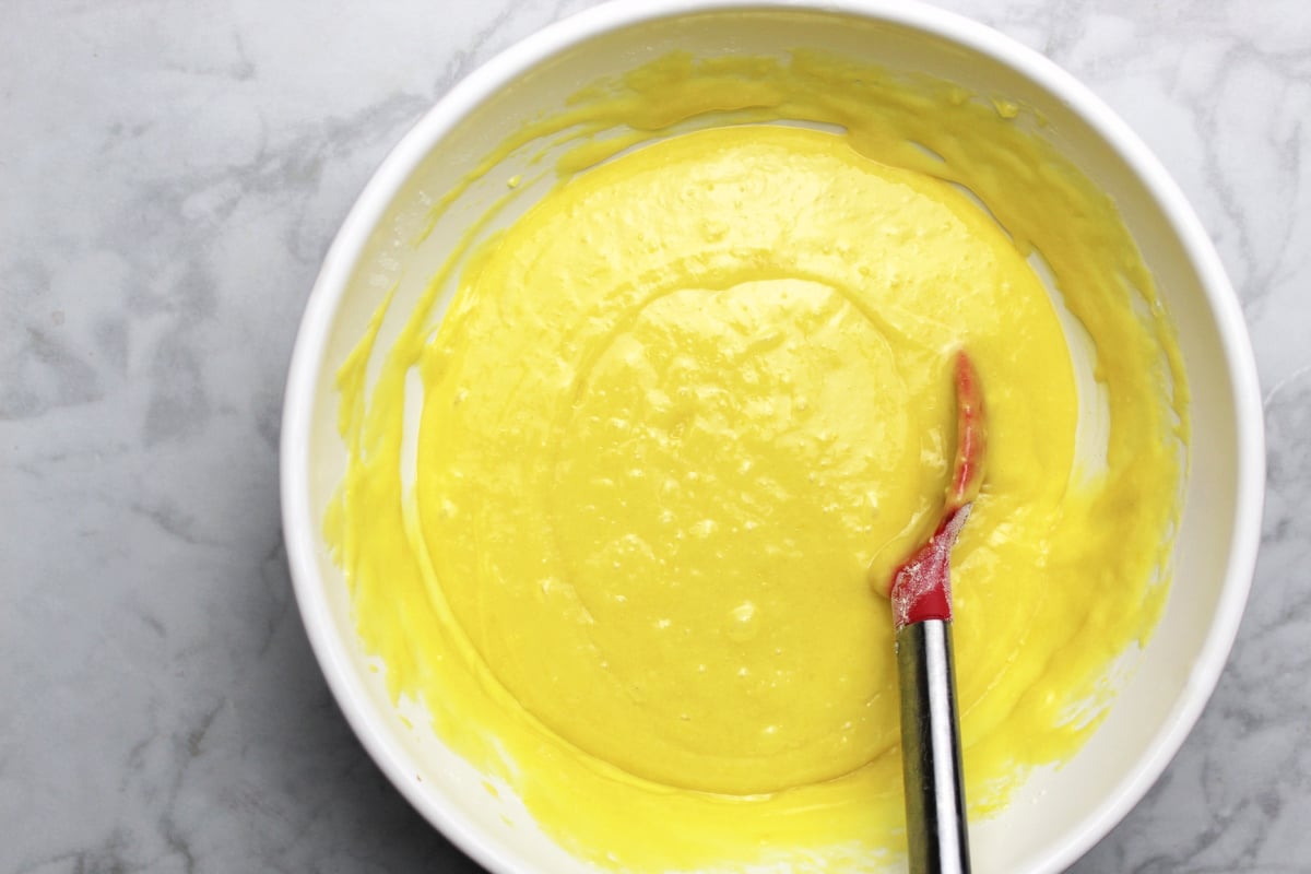 Bright yellow lemon poke batter in a white bowl, with a red spoon used to stir.