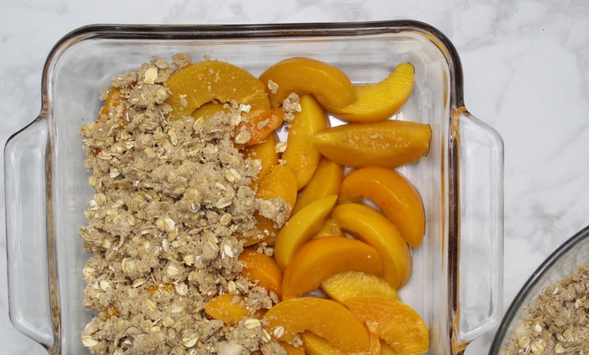 A 9 x 9 pan with the base covered with sliced peaches. Crumble is being spread on top and covers the left half.