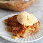 A serving of apple dump cake with a scoop of ice cream on top. In the background is the pan of the apple dump cake.