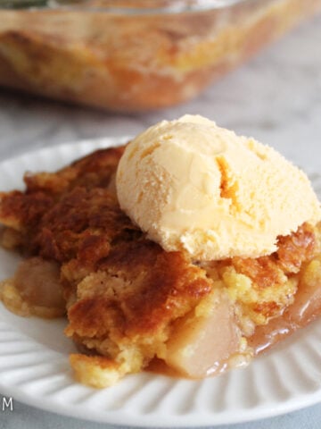 A serving of apple dump cake with a scoop of ice cream on top. In the background is the pan of the apple dump cake.