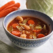 blue bowl of cabbage vegetable soup with carrots and a head of cabbage in the background.