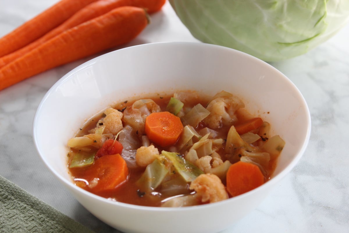 cabbage vegetable soup in a white bowl with cabbage and carrots in the background.