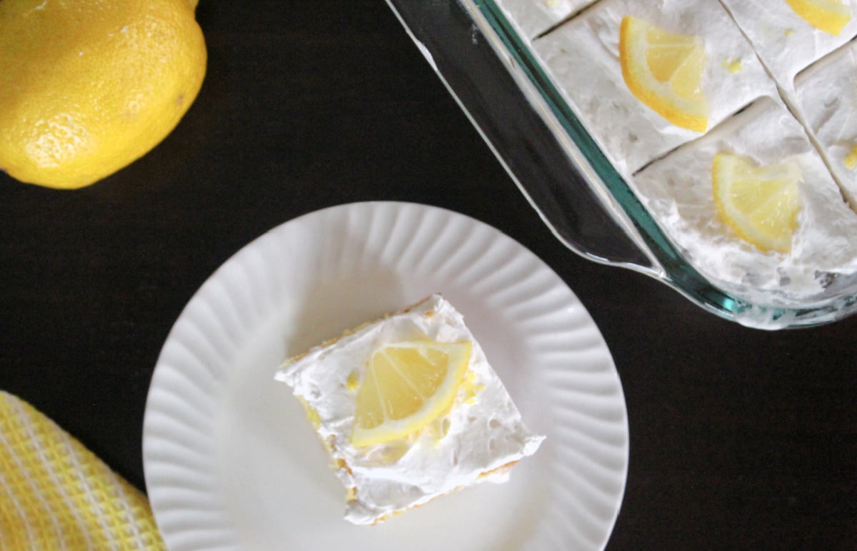 top view of lemon poke cake on a white plate. cake has fluffy frosting and a ¼ slice of lemon as garnish.