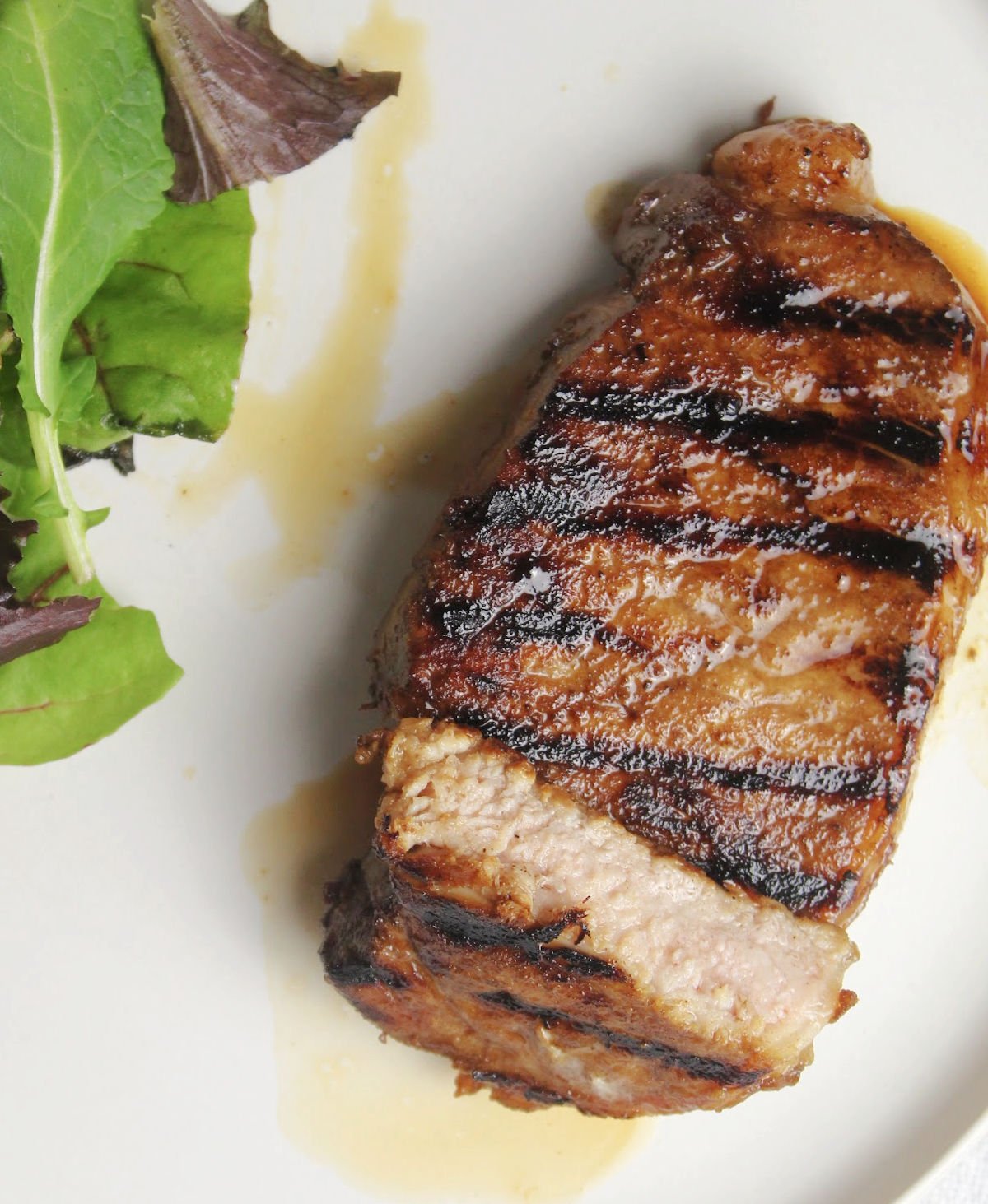 a center cut pork chop with grill marks and juices coming out of the meat on a white plate with a salad beside it.