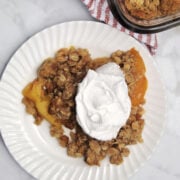 a serving of peach crisp with a dollop of whipped topping. behind the plate is part of the baking dish showing the peach crisp.