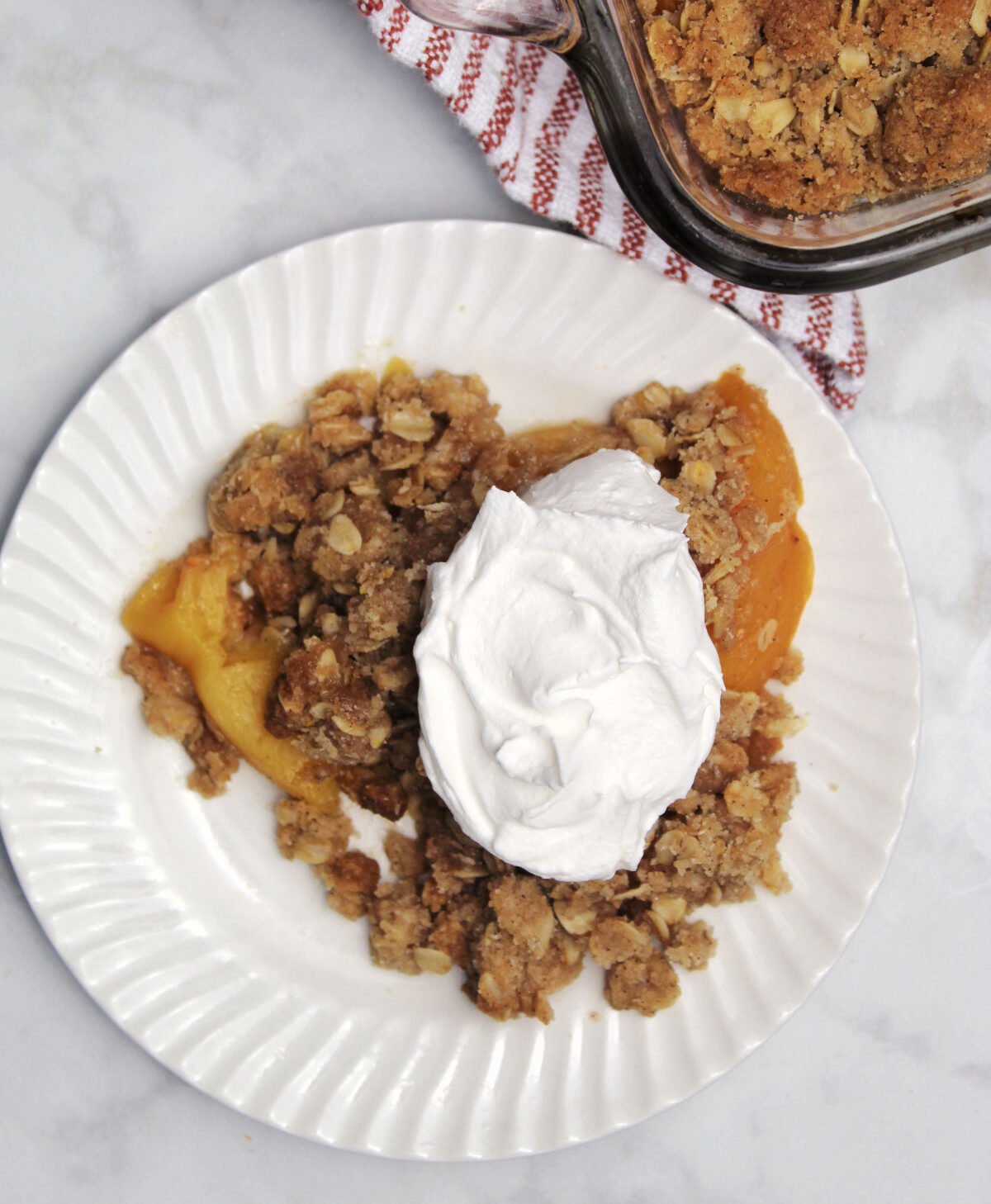 a serving of peach crisp with a dollop of whipped topping. behind the plate is part of the baking dish showing the peach crisp.