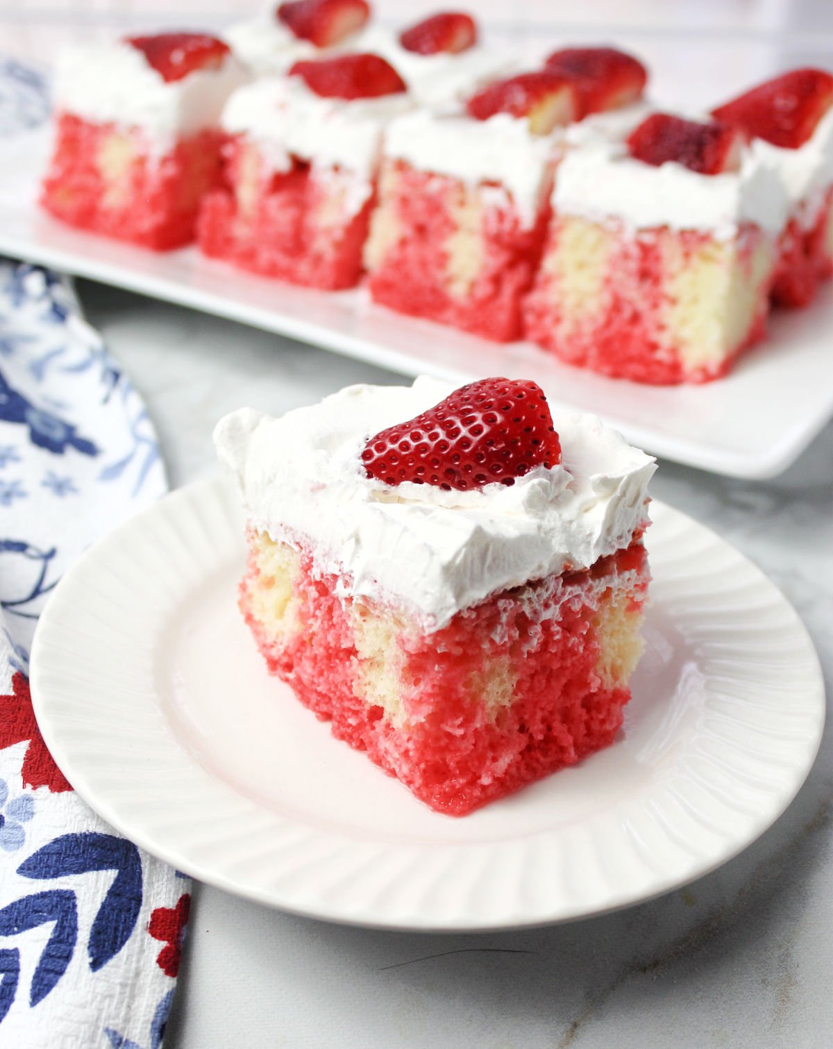 strawberry poke cake on a plate. You can see where the strawberry jello went through the pokes. Covered in whipped topping and a ½ strawberry.