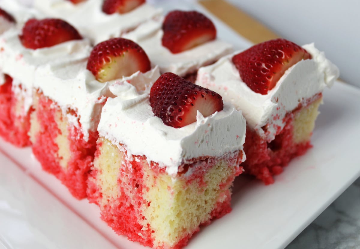 a tray with strawberry poke cake with strawberry jello visible through the cake and then whipped topping with ½ a strawberry on each piece.
