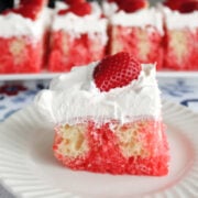 A serving of strawberry poke cake on a white plate with a try of poke cake portions in the background.